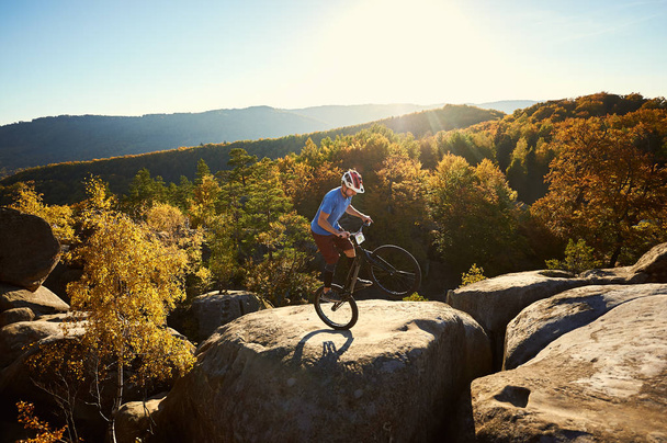 Young cyclist standing on back wheel on trial bicycle, sportsman rider making acrobatic trick on edge of big boulder on top of mountain at sunset - Photo, Image