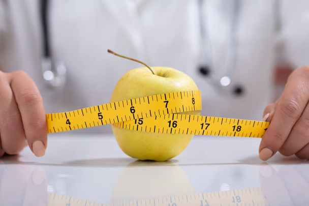 Close-up Of Female Dietitian Measuring The Whole Apple With Measuring Tape On Desk - Φωτογραφία, εικόνα