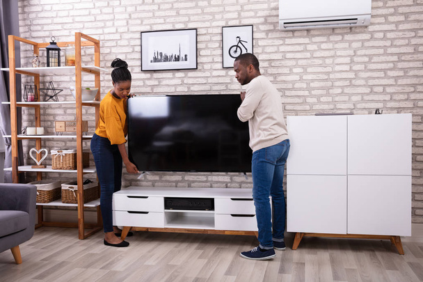 Side View Of Young African Couple Lifting Television At Home - Foto, afbeelding