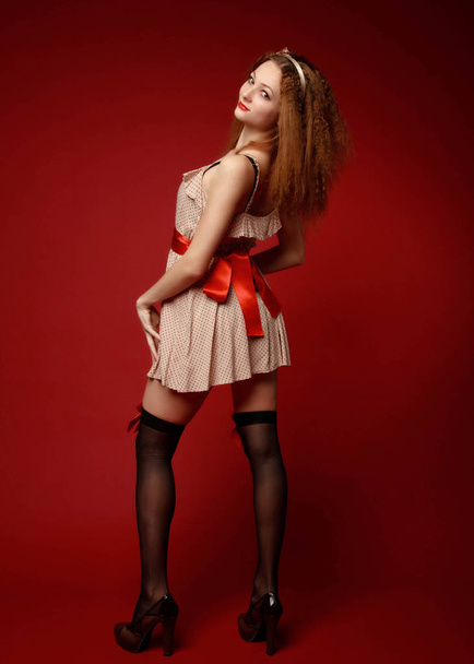Smiling pretty young woman in short dress, red belt, black stockings with red ribbons and high heels stands half-turned against the red background. Pin-up full-growth retro-style portrait - Photo, image