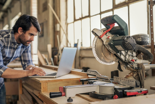Young woodworker working online with a laptop while leaning on bench by a mitre saw in his workshop - Photo, image