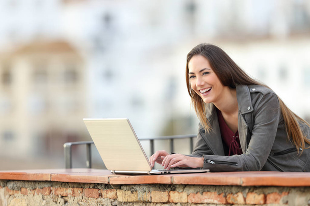 Mulher feliz usando um laptop e olhando para você em uma varanda em uma cidade periferia
 - Foto, Imagem