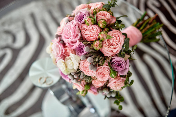 Close up of lush bridal bouquet of pink and purple roses with satin ribbon and two golden rings in blur on glass table background indoors, copy space. Wedding concept  - Zdjęcie, obraz