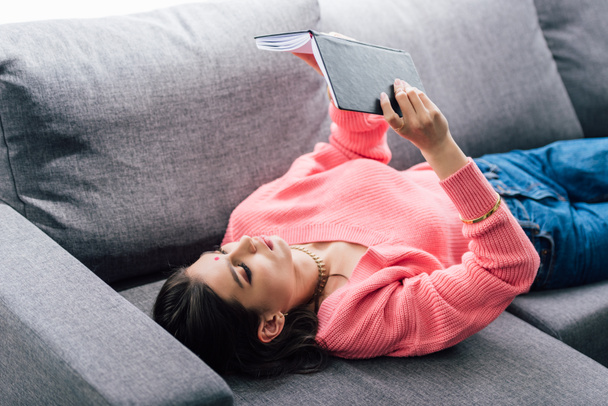 attractive indian student with bindi reading book and lying on sofa - Photo, Image