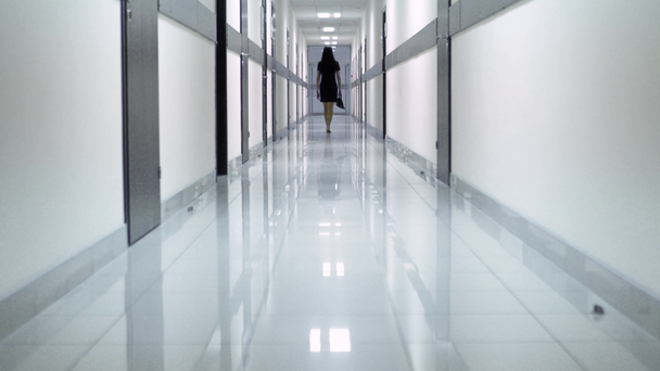 Вид сзади на женщину в черном платье, идущую по коридору отеля. Она держит ботинки в руке
. - Кадры, видео
