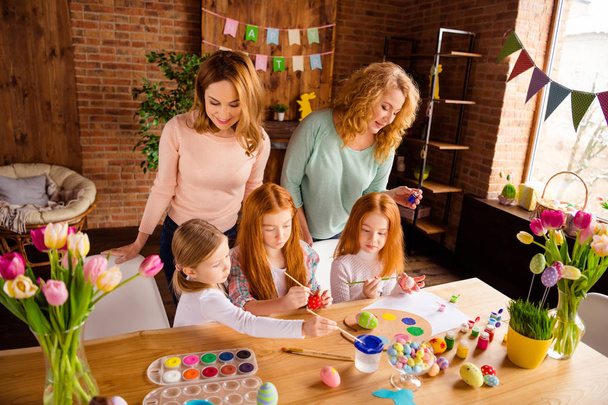 Portrait of two nice attractive cheerful ladies helping assisting two girls doing making decorative things dying eggs in house brick loft industrial interior room indoors - Photo, image