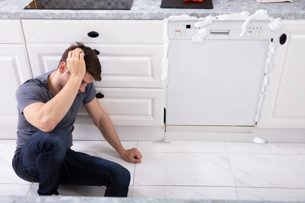 Upset Man Sitting In Front Of Damaged Dishwasher With Foam Coming From It - Фото, изображение