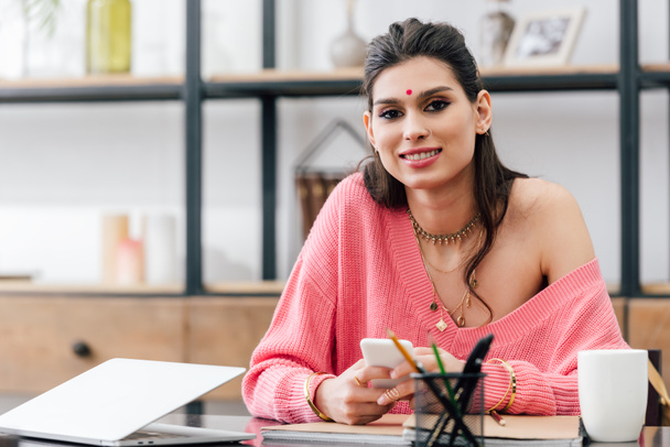 happy indian student with bindi using smartphone at table with laptop - Photo, Image