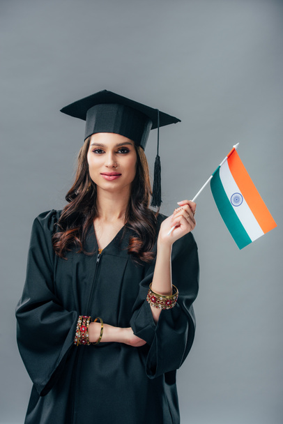 アカデミック ガウンと卒業キャップ グレーに分離されたインドの旗を保持してエレガントなインドの学生 - 写真・画像