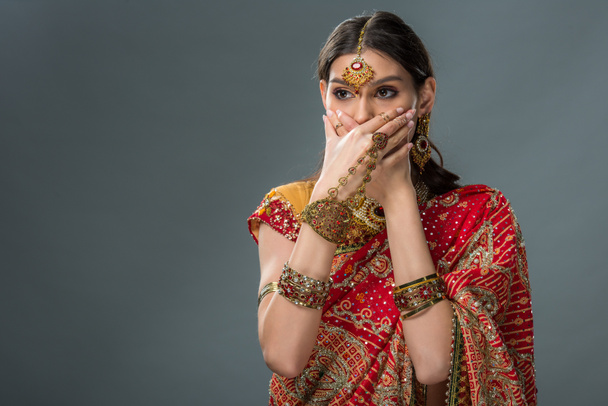όμορφη ινδική γυναίκα που κλείνει το στόμα με τα χέρια, απομονώνονται σε γκρι  - Φωτογραφία, εικόνα