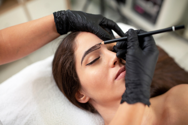 Kosmetikerin bereitet sich auf einen legalen Auftritt von natürlich vollen Augenbrauen auf dem Gesicht einer hübschen jungen Frau vor - Foto, Bild