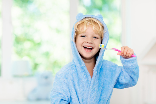 Dişlerini fırçalayan çocuk. Çocukların diş fırçası ve yapıştırıcısı. Mavi banyo bornozu ya da güneşli bir sabahta penceresi olan beyaz banyoda dişlerini fırçalayan havluyla küçük bir oğlan. Çocuklar için diş sağlığı ve çalılık. - Fotoğraf, Görsel