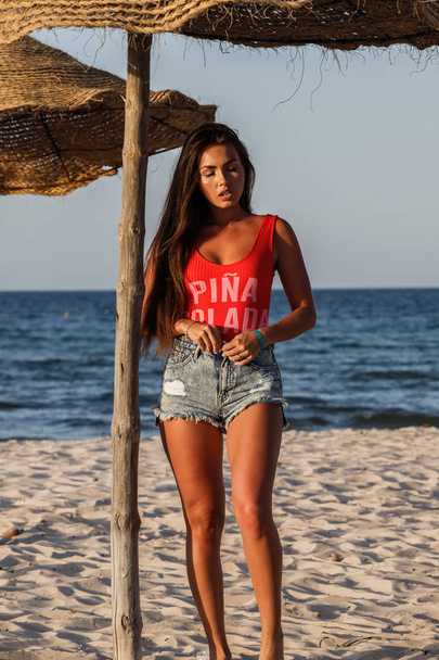 Beautiful brunette model in red swimsuit and blue jeans shorts standing on sandy beach at daytime - 写真・画像