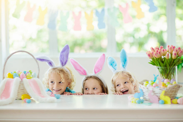 Kids dyeing Easter eggs. Children in bunny ears dye colorful egg for Easter hunt. Home decoration with flowers, basket and rabbit for spring holiday celebration. Little boy and girl decorate home. - Photo, Image