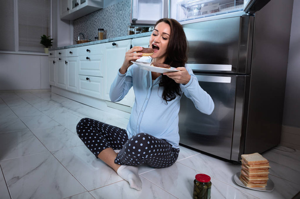 Young Pregnant Woman Sitting On Tiled Floor Eating Donut In Kitchen - Фото, изображение