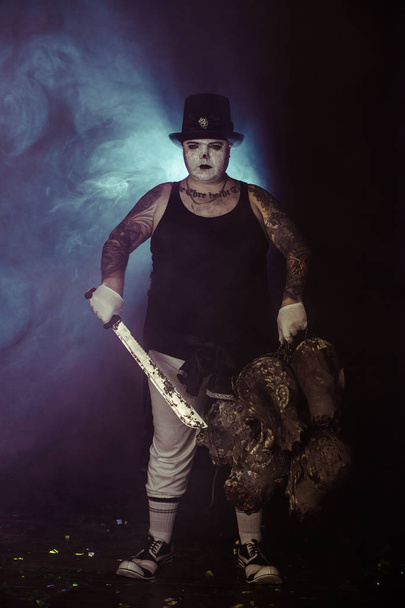 portrait of zombie killer wearing hat with machete and zombie heads posing against dark background - Foto, afbeelding