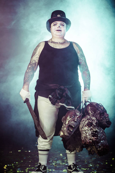 portrait of zombie killer wearing hat with machete and zombie heads posing against dark background - Valokuva, kuva