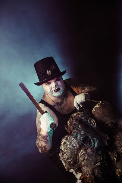 portrait of zombie killer wearing hat with machete and zombie heads posing against dark background - Фото, зображення