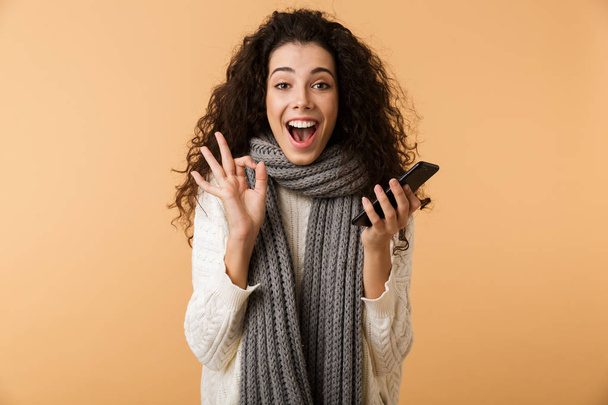 Cheerful young woman wearing winter scarf standing isolated over beige background, holding mobile phone, ok - Fotografie, Obrázek