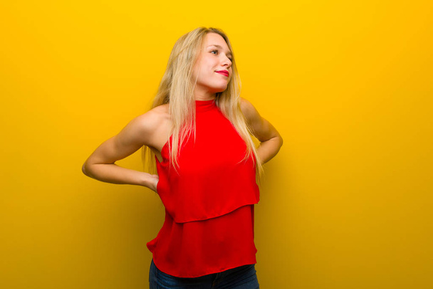 Jong meisje met rode jurk over gele muur lijden aan rugpijn voor een inspanning hebben gedaan - Foto, afbeelding
