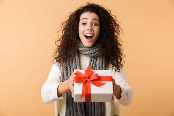 Happy young woman wearing winter scarf standing isolated over beige background, holding present box - Φωτογραφία, εικόνα