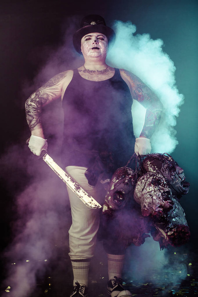 portrait of zombie killer wearing hat with machete and zombie heads posing against dark background - Photo, image