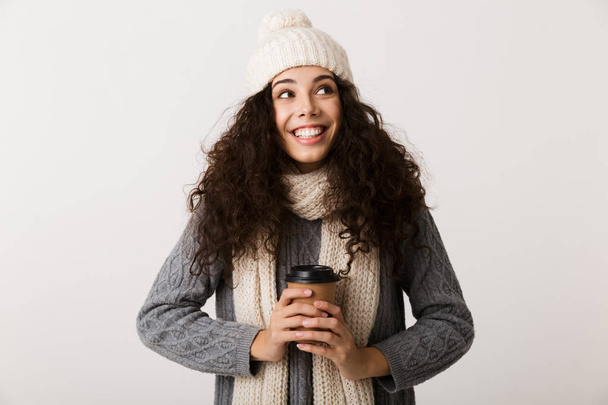 Cheerful young woman wearing winter scarf standing isolated over white background, holding takeaway cup of coffee - Fotó, kép
