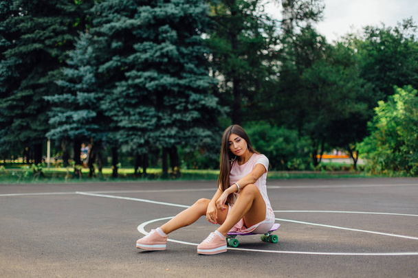 Portrait of a smiling charming brunette female sitting on her skateboard on a basketball court. - 写真・画像
