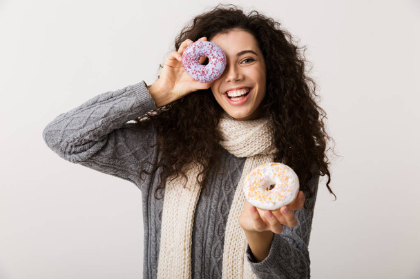Attractive young woman wearing winter scarf showing sweet donuts while standing isolated over white background - Фото, изображение