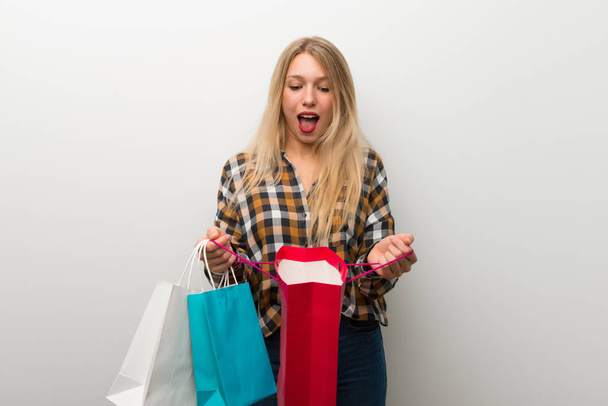 Blonde young girl over white wall surprised while holding a lot of shopping bags - Photo, Image