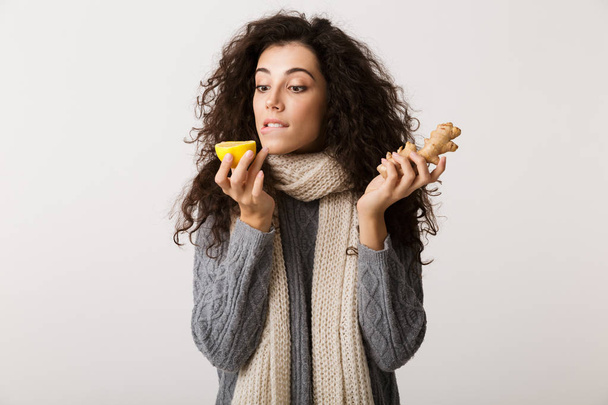 Atractiva mujer joven con bufanda de invierno que muestra jengibre y limón mientras está de pie aislado sobre fondo blanco
 - Foto, Imagen