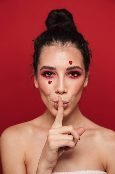 Beauty portrait of an attractive young topless woman wearing makeup standing isolated over red background, showing silence gesture - Foto, Imagen