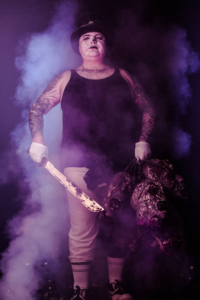 portrait of zombie killer wearing hat with machete and zombie heads posing against dark background - Foto, Imagen