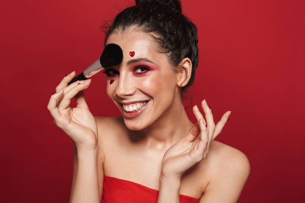 Beauty portrait of an attractive young topless woman wearing makeup standing isolated over red background, posing, holding a makeup brush - 写真・画像