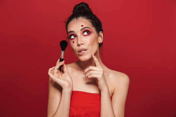 Beauty portrait of an attractive young topless woman wearing makeup standing isolated over red background, posing, holding a makeup brush - Foto, afbeelding