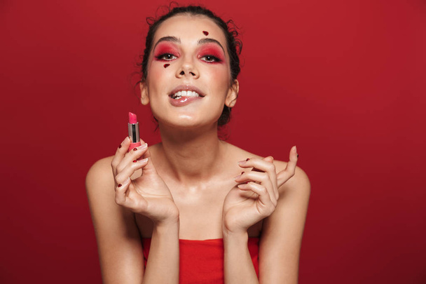 Beauty portrait of an attractive young topless woman wearing makeup standing isolated over red background, posing, holding a lipstick - Foto, Imagen