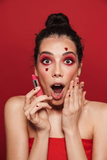 Beauty portrait of an attractive young topless woman wearing makeup standing isolated over red background, posing, holding a lipstick - 写真・画像