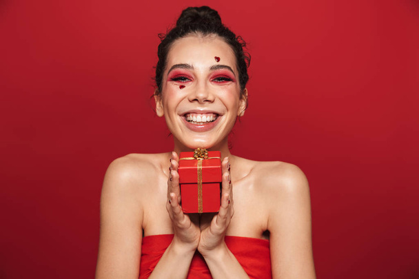Beauty portrait of an attractive young topless woman wearing makeup standing isolated over red background, posing, holding a gift box - Photo, Image