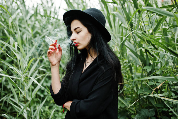 Sensual smoker girl all in black, red lips and hat. Goth dramatic woman smoking on common reed. - Foto, imagen