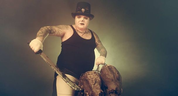 portrait of zombie killer wearing hat with machete and zombie heads posing against dark background - Foto, immagini