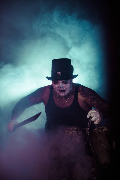 portrait of zombie killer wearing hat with machete and zombie heads posing against dark background - Zdjęcie, obraz