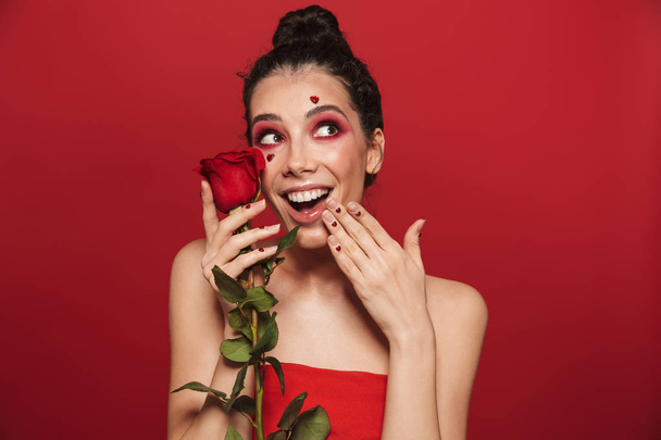 Beauty portrait of an attractive young topless woman wearing makeup standing isolated over red background, posing with a red rose - Photo, Image