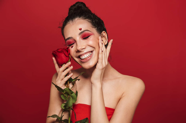 Beauty portrait of an attractive young topless woman wearing makeup standing isolated over red background, posing with a red rose - Φωτογραφία, εικόνα