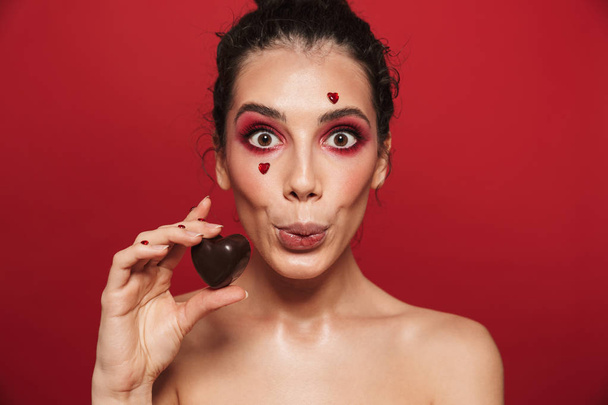 Beauty portrait of an attractive young topless woman wearing makeup standing isolated over red background, holding heart shaped chocolate candy - Φωτογραφία, εικόνα