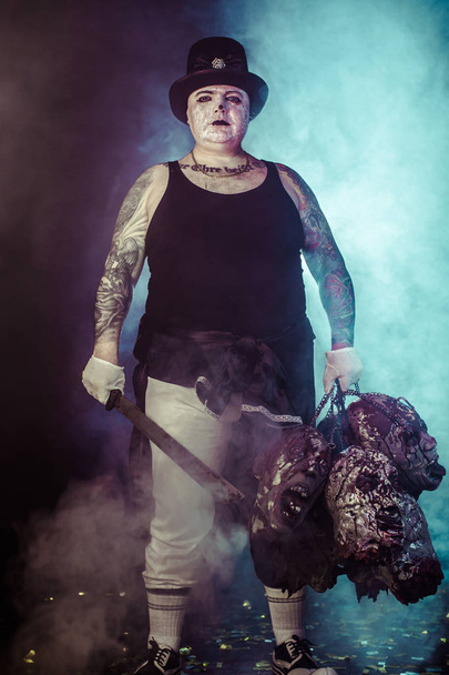 portrait of zombie killer wearing hat with machete and zombie heads posing against dark background - Fotó, kép