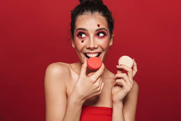 Beauty portrait of an attractive young topless woman wearing makeup standing isolated over red background, eating macaroons - Фото, зображення