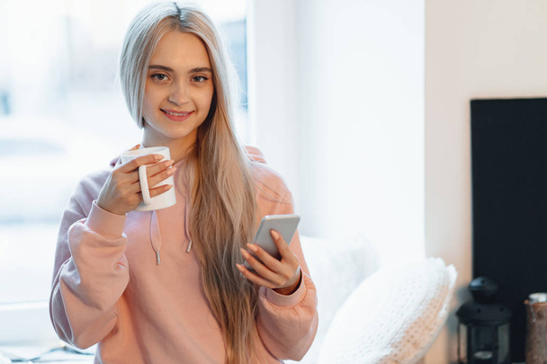 Mooi meisje zittend op de vensterbank met een kopje koffie, thee en smartphone in handen. Ze heeft lang blond haar, glimlach en kijken naar haar telefoon. Het dragen van roze, bleke parka. - Foto, afbeelding