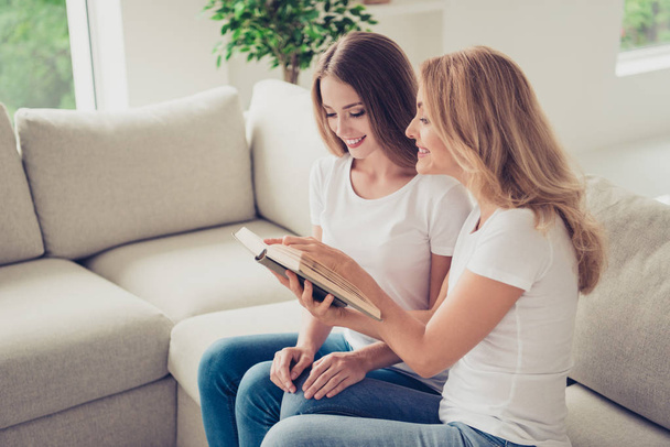 Close up photo of two people mum and teen daughter lovely look to open book holding hands arms adventure novel showing favorite moment wear white t-shirts jeans sit on comfy sofa - Foto, afbeelding