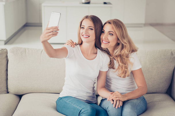 Two nice cute attractive charming lovely winsome lovable fascinating tender cheerful ladies sitting on divan using gadget making taking selfie in light white interior room indoors - Fotografie, Obrázek