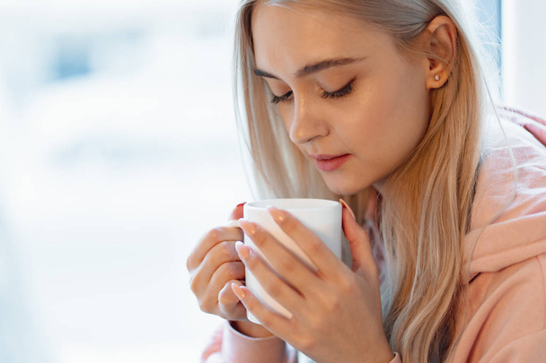 A pretty smiling girl enjoying a cup of coffee or milk while sitting by the blurred window glass. Wearing a casual pink, pale parka. Close up white mug in woman hands near lips. - Photo, Image